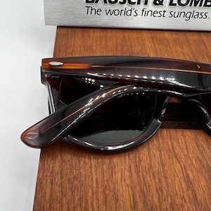 Puede incluir: Gafas de sol marrones oscuras con montura rectangular y lentes oscuras. Las patillas son de color marr&oacute;n claro con una raya naranja. El texto "The world's finest sunglass" es visible encima de las gafas de sol.