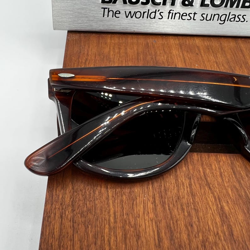 Puede incluir: Gafas de sol marrones oscuras con montura rectangular y lentes oscuras. Las patillas son de color marr&oacute;n claro con una raya naranja. El texto "The world's finest sunglass" es visible encima de las gafas de sol.