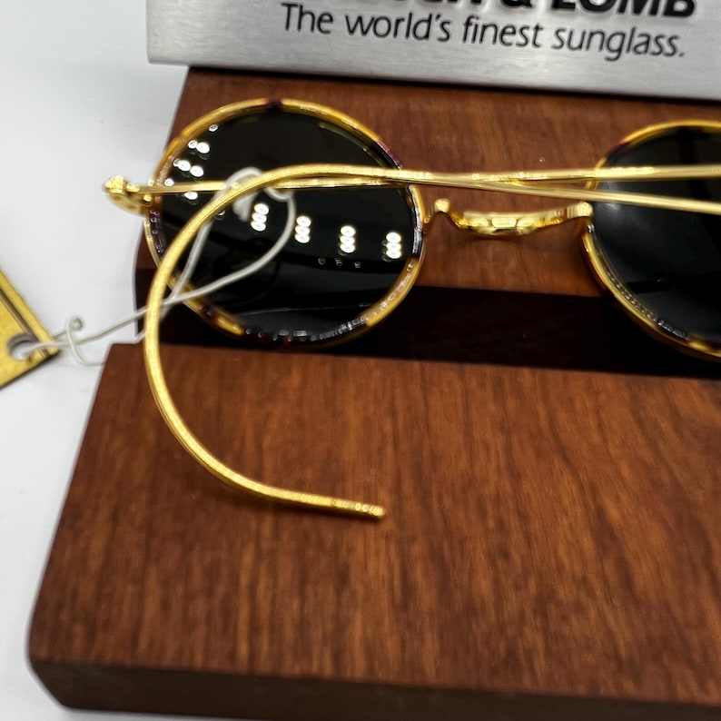 K&ouml;nnte beinhalten: Runde Sonnenbrille mit goldenem Rahmen und dunklen Gl&auml;sern, pr&auml;sentiert auf einer Holzoberfl&auml;che. Der Text "The world's finest sunglass" ist &uuml;ber der Sonnenbrille zu sehen. Die Rahmen haben ein klassisches, elegantes Design.