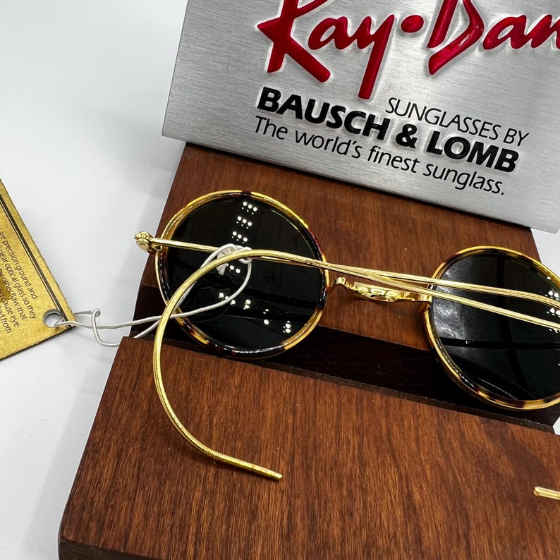 K&ouml;nnte beinhalten: Runde Sonnenbrille mit goldenem Rahmen und dunklen Gl&auml;sern. Die Gl&auml;ser haben ein Schildpattmuster an den R&auml;ndern. Im Hintergrund ist der Text "Kay Dan" und "BAUSCH & LOMB" zu sehen.
