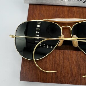 May include: Gold-rimmed sunglasses with dark lenses. The sunglasses have a classic aviator style with a double bridge and curved temples. The lenses are a dark, reflective color, and the frame is a shiny gold. The sunglasses are resting on a wooden surface.