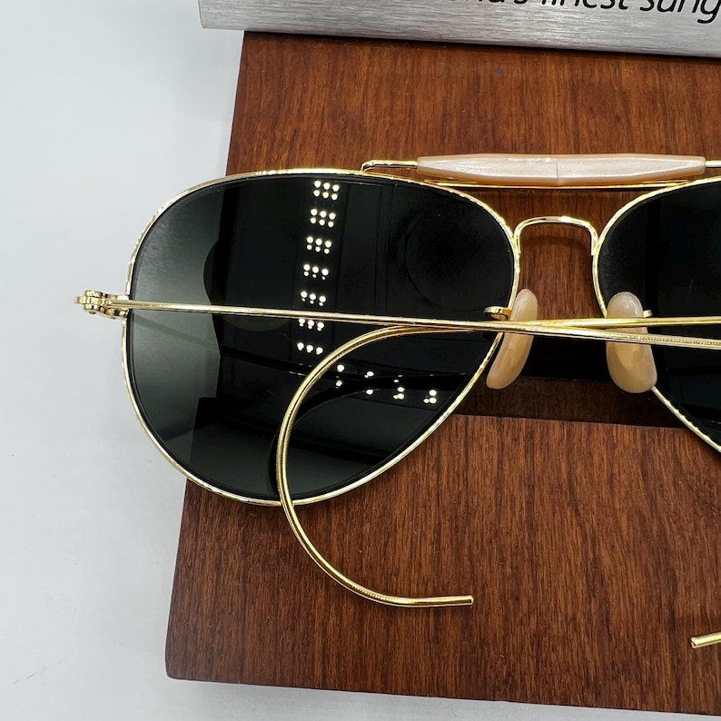 May include: Gold-rimmed sunglasses with dark lenses. The sunglasses have a classic aviator style with a double bridge and curved temples. The lenses are a dark, reflective color, and the frame is a shiny gold. The sunglasses are resting on a wooden surface.