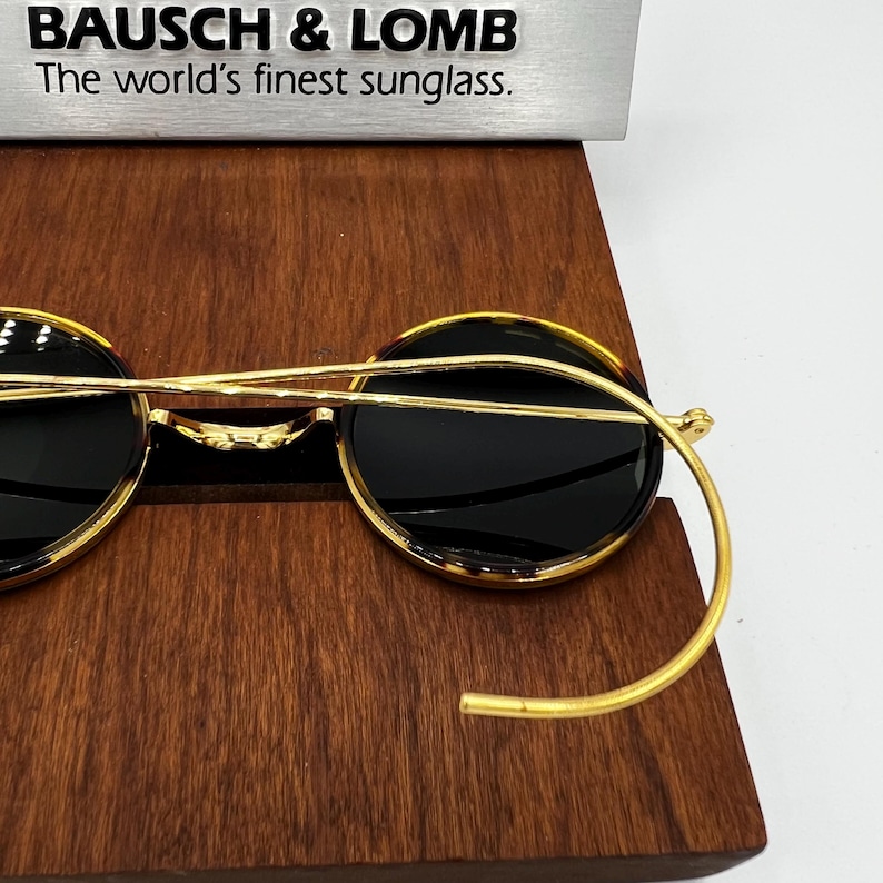 K&ouml;nnte beinhalten: Goldrand-Sonnenbrille mit dunklen Gl&auml;sern auf einer Holzoberfl&auml;che. Die Sonnenbrille hat eine runde Form und ein markantes gebogenes B&uuml;geldesign. Der Text "BAUSCH & LOMB The world's finest sunglass." ist sichtbar.