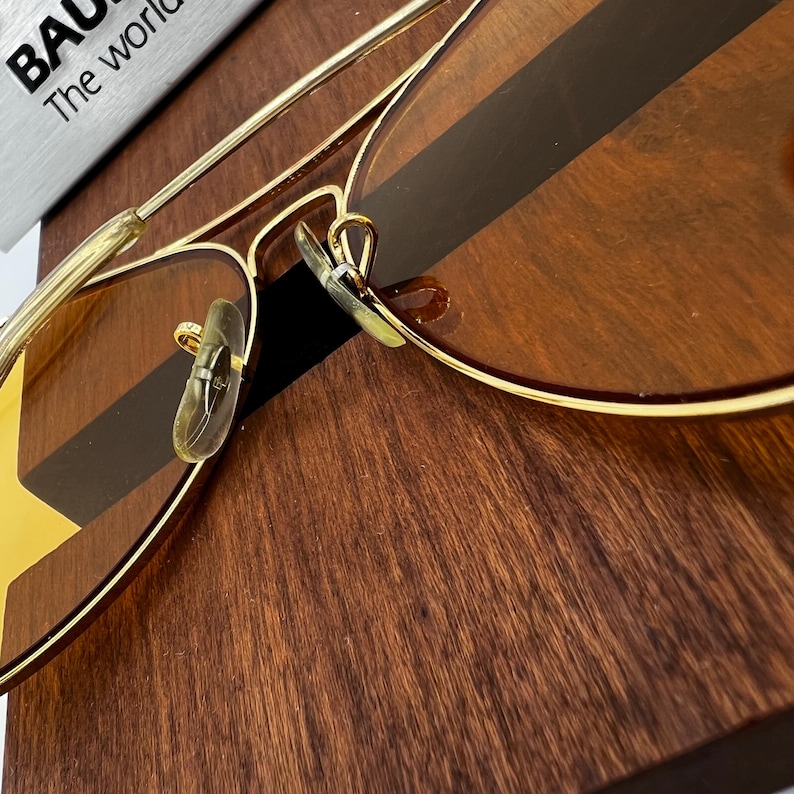 Puede incluir: Gafas de sol tipo aviador de metal dorado con lentes tintadas en &aacute;mbar y dise&ntilde;o de doble puente. Las almohadillas nasales son transl&uacute;cidas y la montura incluye patillas de alambre fino. Una etiqueta parcial muestra BAUSCH y The world.