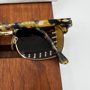 May include: Close-up of sunglasses with a tortoiseshell pattern on the arms and a gold-colored frame. The lenses are dark, and the sunglasses are resting on a wooden surface. The design is classic and stylish.