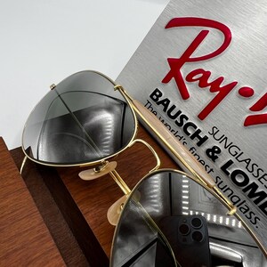 May include: Gold-rimmed sunglasses with dark lenses rest on a wooden surface. The sunglasses are positioned in front of a silver sign with the red Ray-Ban logo and the text "BAUSCH & LOMB SUNGLASSES The world's finest sunglasses."
