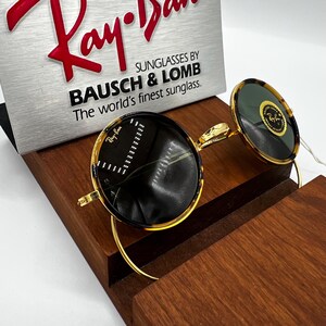 K&ouml;nnte beinhalten: Runde Sonnenbrille mit Schildpatt-Fassung und goldfarbenen B&uuml;geln. Die Gl&auml;ser sind dunkel, und die Sonnenbrille wird auf einem Holzst&auml;nder pr&auml;sentiert. Der Text "Ray-Ban" ist auf dem Glas sichtbar.