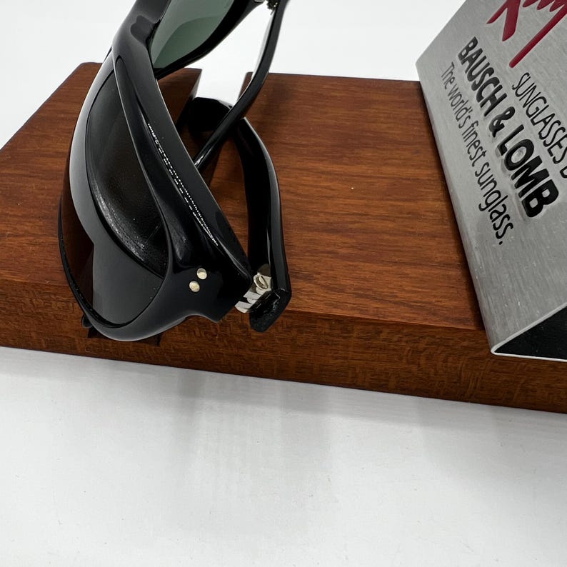 May include: Black sunglasses with a sleek, curved design rest on a wooden display. The sunglasses have dark lenses and a glossy finish. The text on the display reads "BAUSCH & LOMB Sunglasses, The world's finest sunglass."