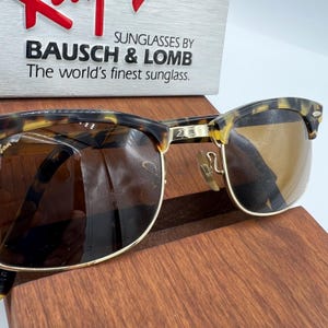 May include: Sunglasses by Bausch & Lomb with a tortoiseshell frame and brown lenses. The sunglasses have a gold-colored metal frame and are displayed on a wooden surface. The text "The world's finest sunglass" is visible.