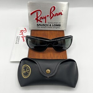 May include: Black Ray-Ban sunglasses with a matching black case and a silver sign with the Ray-Ban logo. The sunglasses are displayed on a wooden surface, with a booklet featuring the Ray-Ban logo.