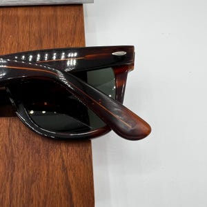 Puede incluir: Gafas de sol marrones oscuras con un patr&oacute;n de carey. Las gafas de sol tienen una montura rectangular cl&aacute;sica y lentes oscuras. La patilla de las gafas de sol est&aacute; extendida, revelando la bisagra y el interior de la montura.