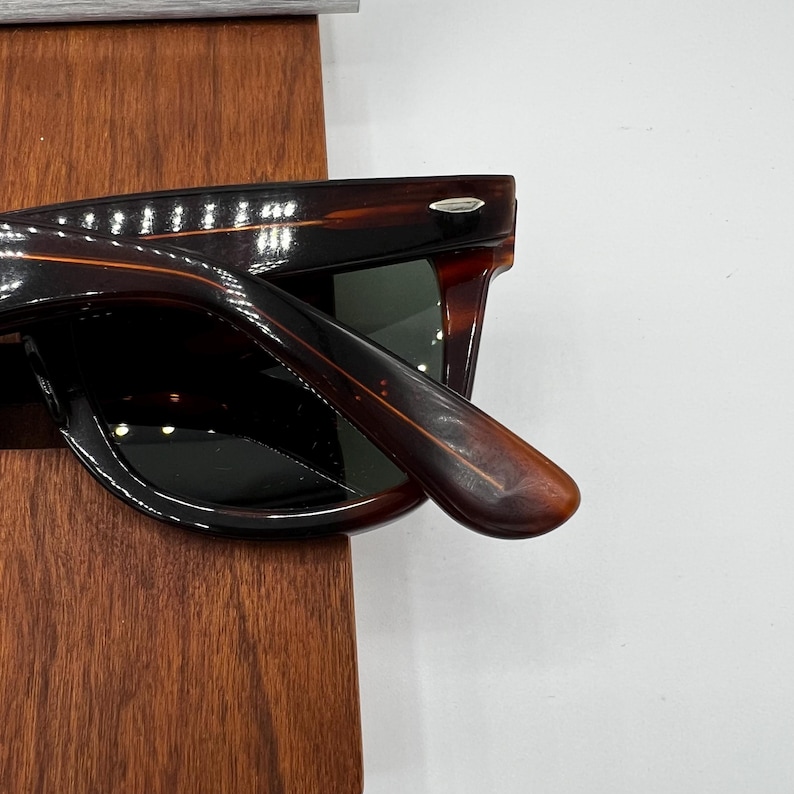 Puede incluir: Gafas de sol marrones oscuras con un patr&oacute;n de carey. Las gafas de sol tienen una montura rectangular cl&aacute;sica y lentes oscuras. La patilla de las gafas de sol est&aacute; extendida, revelando la bisagra y el interior de la montura.