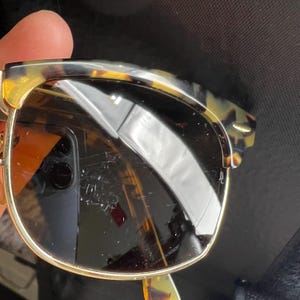 May include: Sunglasses with a tortoiseshell pattern on the top frame and temples. The lenses are a dark brown color, and the frames have a gold-colored metal rim. The sunglasses are held up by a hand.