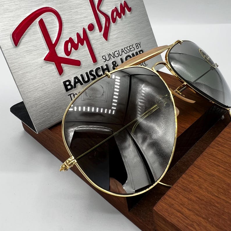 May include: Gold-rimmed aviator sunglasses with dark lenses. The sunglasses are displayed on a wooden stand next to a silver sign with the red Ray-Ban logo and text. The lenses reflect the surrounding environment.
