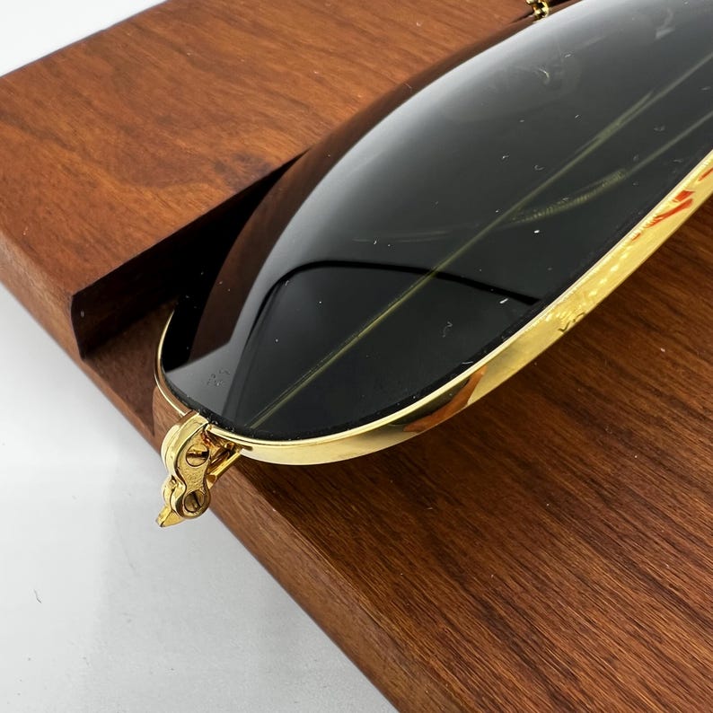 Pode incluir: Close-up de &oacute;culos de sol com arma&ccedil;&atilde;o dourada e lentes escuras. Os &oacute;culos de sol est&atilde;o apoiados em uma superf&iacute;cie de madeira. A arma&ccedil;&atilde;o dourada tem um design detalhado perto das dobradi&ccedil;as.