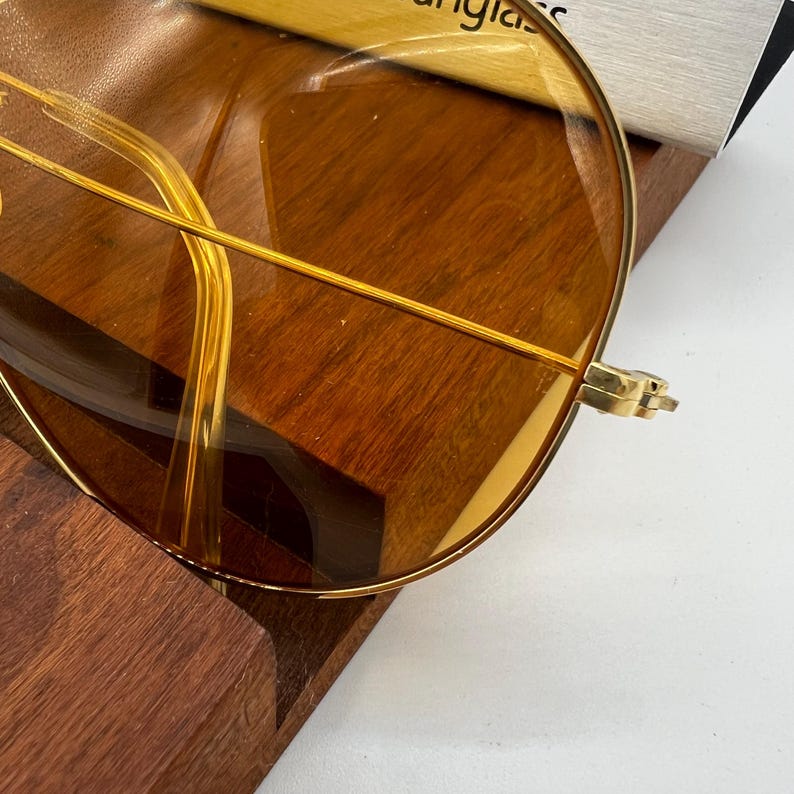Puede incluir: Gafas de sol con montura fina de metal dorado y lentes grandes, redondas y tintadas en color &aacute;mbar. El dise&ntilde;o incluye un puente dorado delgado que conecta ambas lentes, con varillas doradas visibles a trav&eacute;s del cristal transparente.
