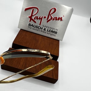 May include: Gold-rimmed sunglasses with green lenses, resting on a wooden display stand. A silver sign with the red "Ray-Ban" logo and the text "BAUSCH & LOMB SUNGLASSES BY The world's finest sunglass." is in the background.