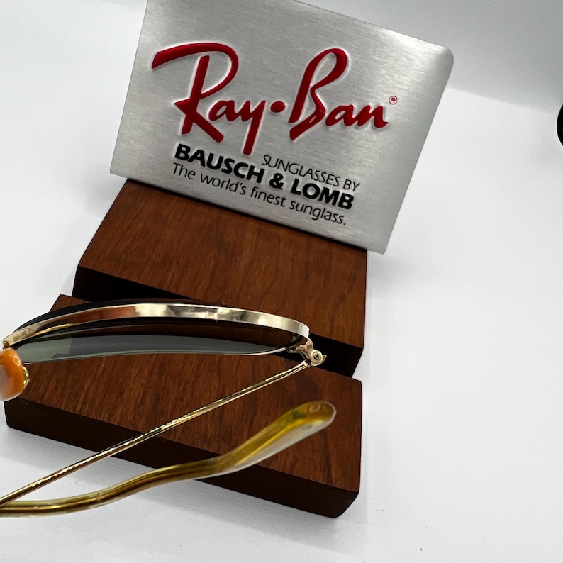 May include: Gold-rimmed sunglasses with green lenses, resting on a wooden display stand. A silver sign with the red "Ray-Ban" logo and the text "BAUSCH & LOMB SUNGLASSES BY The world's finest sunglass." is in the background.