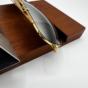 May include: Gold-rimmed sunglasses with dark lenses rest on a wooden display stand. The sunglasses have a sleek, modern design with a gold frame and dark lenses. The wooden stand is a rich, dark brown color.
