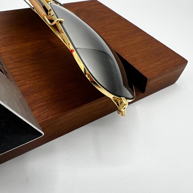 May include: Gold-rimmed sunglasses with dark lenses rest on a wooden display stand. The sunglasses have a sleek, modern design with a gold frame and dark lenses. The wooden stand is a rich, dark brown color.
