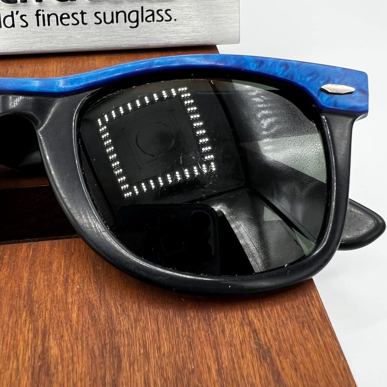 Op de afbeelding: Zwarte zonnebril met een blauw gemarmerd bovenframe. De lenzen zijn donker en reflecteren de omgeving. De zonnebril wordt gepresenteerd op een houten oppervlak. De tekst "d's finest sunglass" is zichtbaar op de achtergrond.