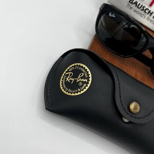 May include: Black Ray-Ban sunglasses with a matching black case. The case has a gold emblem with the Ray-Ban logo and text. The sunglasses are resting on a wooden surface, with a book in the background.