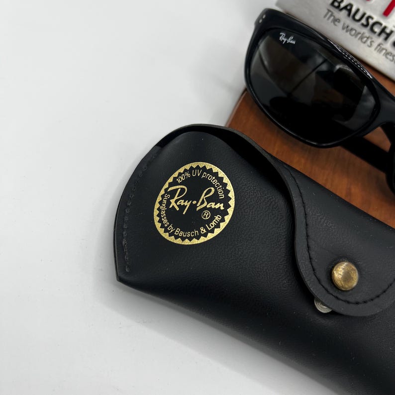 May include: Black Ray-Ban sunglasses with a matching black case. The case has a gold emblem with the Ray-Ban logo and text. The sunglasses are resting on a wooden surface, with a book in the background.