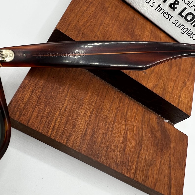 May include: Close-up of a pair of brown tortoiseshell sunglasses resting on a wooden display stand. The sunglasses have a classic design with dark lenses and a thick frame. The text on the frame reads "RAY-BAN".
