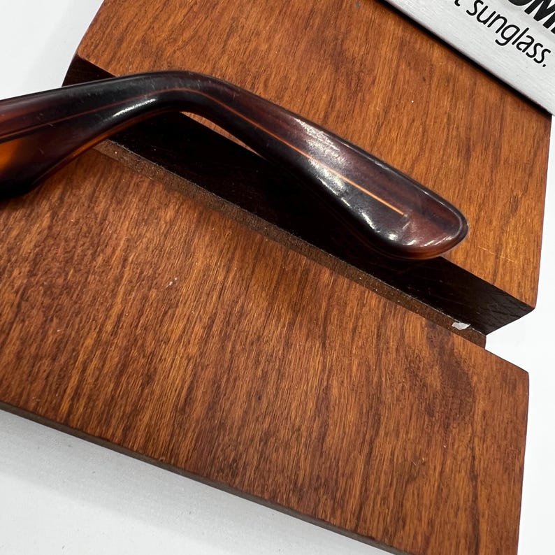 May include: Close-up of a brown and tortoise shell sunglasses arm resting on a wooden sunglasses case. The case is made of dark brown wood with a visible wood grain. The sunglasses arm is curved and has a glossy finish.