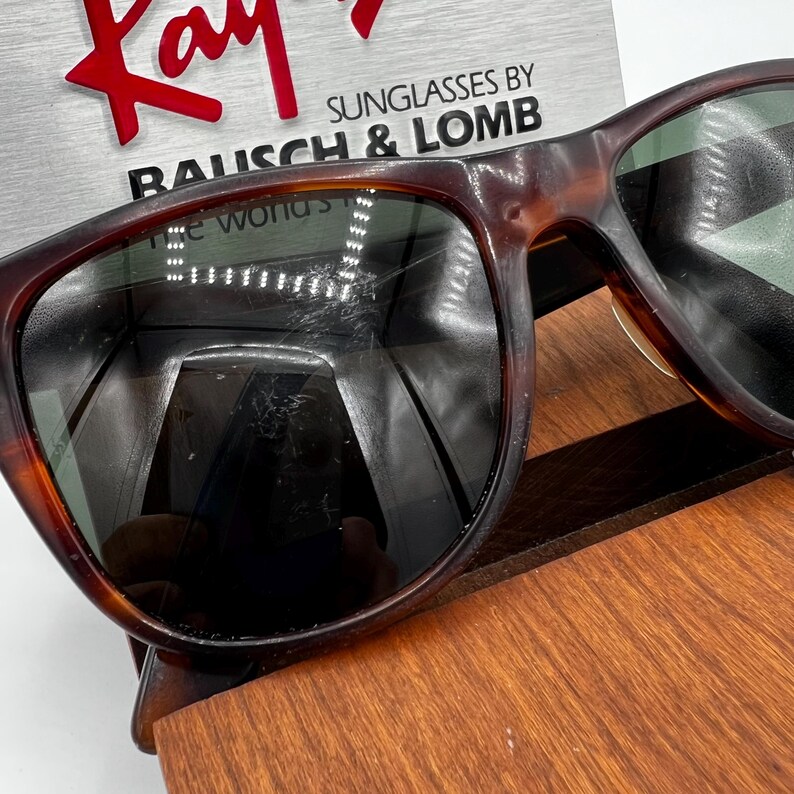 May include: Brown tortoiseshell sunglasses with green lenses. The sunglasses are displayed on a wooden surface. The brand name "Ray-Ban" is visible in red, along with "Sunglasses by Bausch & Lomb".