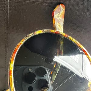 以下が含まれることがあります： 装飾的なハンドルが付いた丸い手鏡。鏡のフレームとハンドルは、黄色、オレンジ、赤の色合いのカラフルな模様のデザインです。鏡の表面は暗くぼやけた画像を反射しています。
