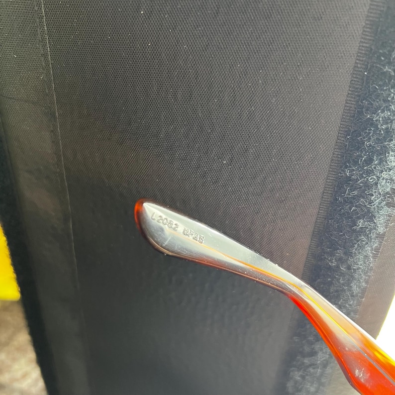Puede incluir: Primer plano de una parte de gafas. La patilla de la montura es plateada con el texto "L2052 62P18" impreso. La patilla hace la transici&oacute;n a un color rojo-naranja transl&uacute;cido. El fondo es negro.