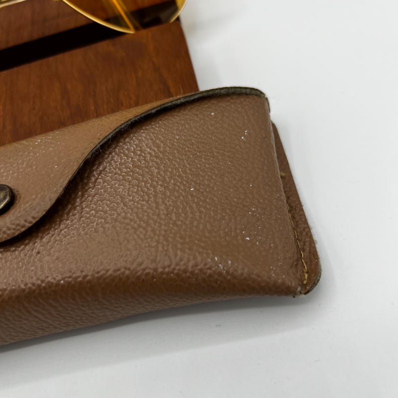 Puede incluir: Un estuche para gafas de color tostado con textura, cierre de solapa y un bot&oacute;n de presi&oacute;n de metal oscuro, con un acabado de superficie granulada. Se ve una parte de unas gafas con montura dorada y lentes amarillas en el borde superior.
