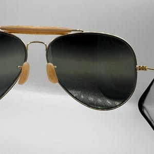 May include: Gold-framed aviator sunglasses with dark lenses. The sunglasses have a double bridge and light-colored nose pads. The top bar is a light brown color. The lenses are a dark green color.