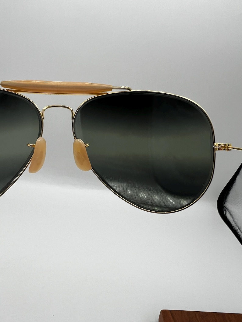 May include: Gold-framed aviator sunglasses with dark lenses. The sunglasses have a double bridge and light-colored nose pads. The top bar is a light brown color. The lenses are a dark green color.