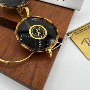 K&ouml;nnte beinhalten: Nahaufnahme einer Ray-Ban-Sonnenbrille mit Schildpattrahmen und dunklen Gl&auml;sern. Die goldfarbenen Metallb&uuml;gel und der Steg bilden einen Kontrast zu den dunklen Gl&auml;sern. Die Sonnenbrille liegt auf einer Holzoberfl&auml;che, im Hintergrund ist ein Etikett zu sehen.