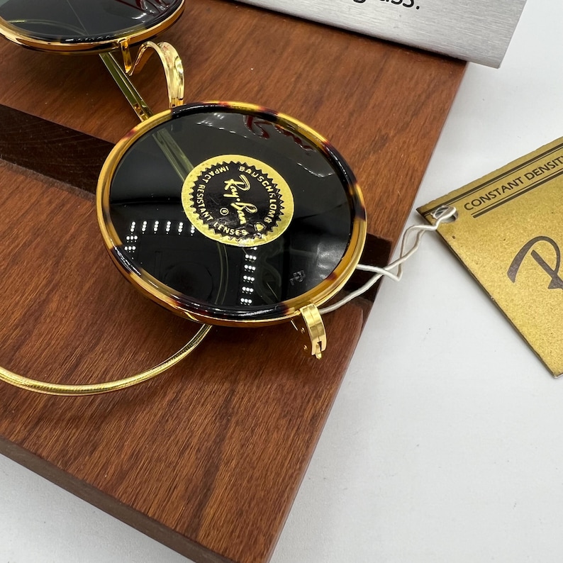 K&ouml;nnte beinhalten: Nahaufnahme einer Ray-Ban-Sonnenbrille mit Schildpattrahmen und dunklen Gl&auml;sern. Die goldfarbenen Metallb&uuml;gel und der Steg bilden einen Kontrast zu den dunklen Gl&auml;sern. Die Sonnenbrille liegt auf einer Holzoberfl&auml;che, im Hintergrund ist ein Etikett zu sehen.