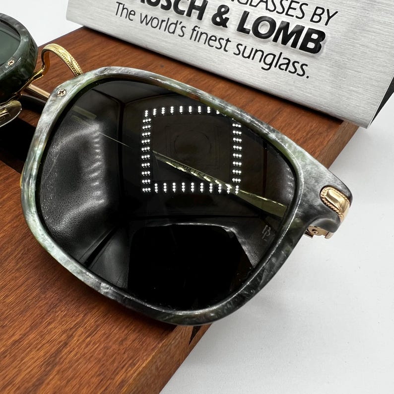 Pu&ograve; includere: Primo piano di occhiali da sole con montatura verde e grigia marmorizzata e lenti scure. Gli occhiali da sole hanno dettagli color oro e sono appoggiati su una superficie di legno. Il testo sul cartello recita "Bausch & Lomb The world's finest sunglass."