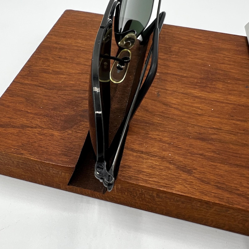 K&ouml;nnte beinhalten: Schwarze Sonnenbrille mit gr&uuml;nen Gl&auml;sern liegt auf einem Holzst&auml;nder. Die Sonnenbrille hat ein klassisches Design mit einem schlanken Rahmen. Der St&auml;nder ist aus dunklem Holz gefertigt und hat ein schlichtes, minimalistisches Design.