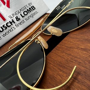 May include: Close-up of sunglasses with black lenses and gold-colored frames. The sunglasses have a classic design with a double bridge and a curved brow bar. The text "SUNGLASSES BY BAUSCH & LOMB" is visible in the background.