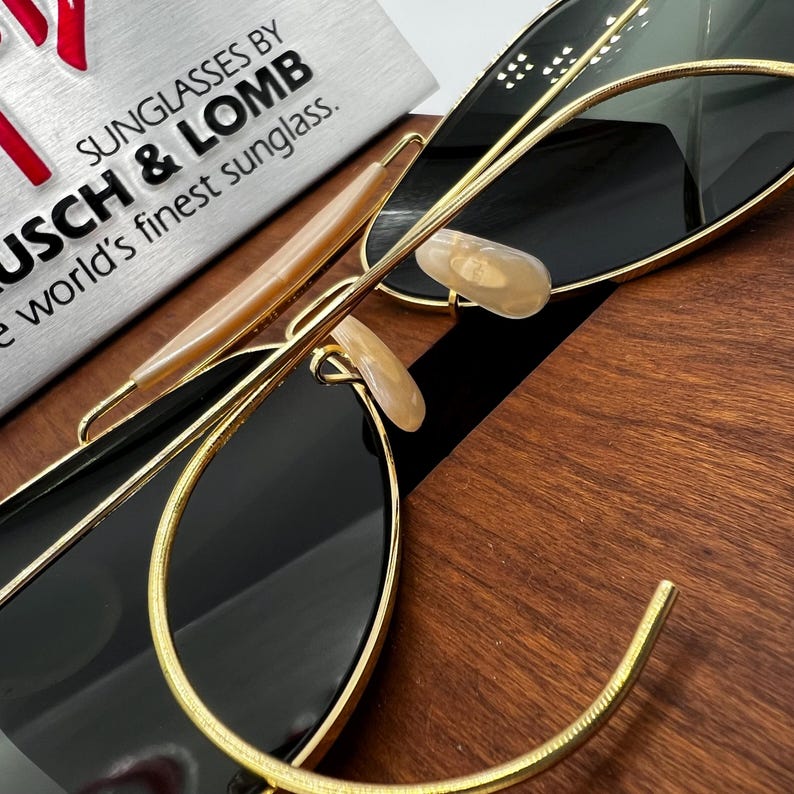 May include: Close-up of sunglasses with black lenses and gold-colored frames. The sunglasses have a classic design with a double bridge and a curved brow bar. The text "SUNGLASSES BY BAUSCH & LOMB" is visible in the background.