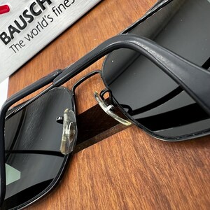 K&ouml;nnte beinhalten: Schwarze Sonnenbrille mit rechteckigem Rahmen und dunklen Gl&auml;sern. Die Sonnenbrille hat einen Metallrahmen und transparente Nasenpads. Der Text "BAUSCH The world's finest" ist im Hintergrund zu sehen.
