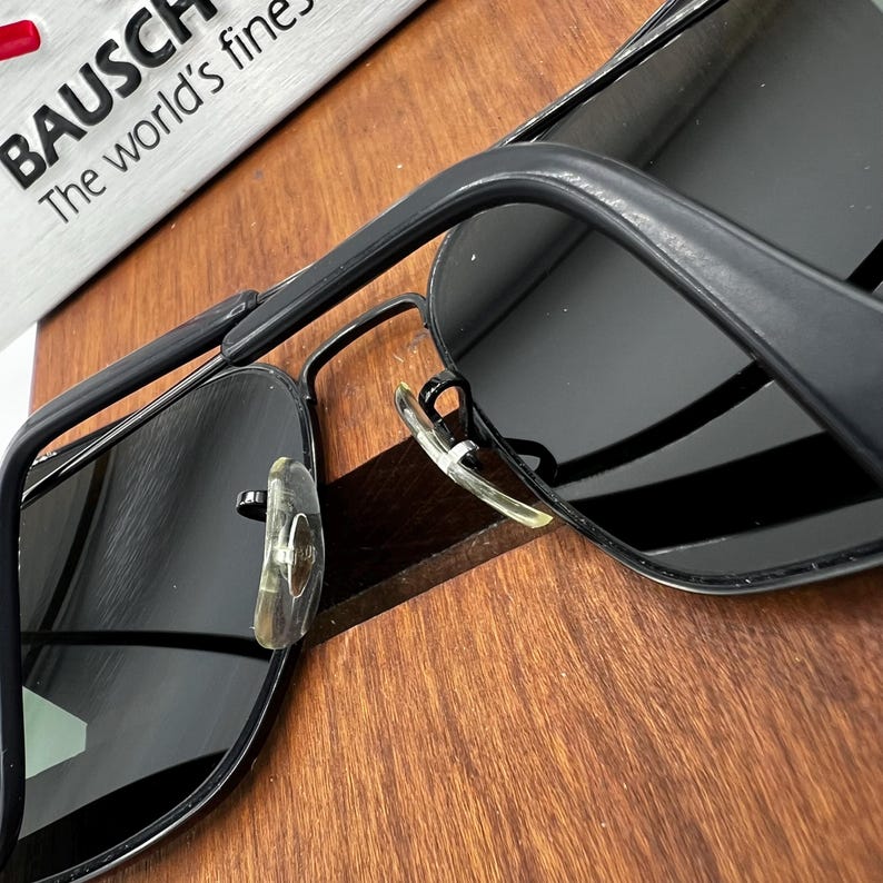 K&ouml;nnte beinhalten: Schwarze Sonnenbrille mit rechteckigem Rahmen und dunklen Gl&auml;sern. Die Sonnenbrille hat einen Metallrahmen und transparente Nasenpads. Der Text "BAUSCH The world's finest" ist im Hintergrund zu sehen.