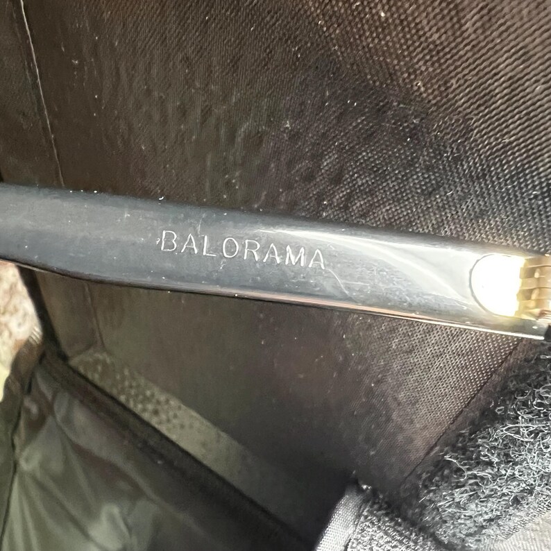 May include: A silver-colored metal object with the word "BALORAMA" engraved on it. The object is partially visible, with a gold-colored, textured section at the end. The item is set against a dark background, possibly inside a case.