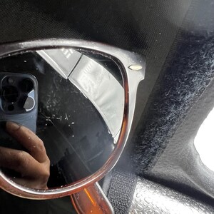 May include: Brown tortoiseshell sunglasses with dark lenses. The sunglasses are reflected in a dark surface, with a phone and a hand visible in the reflection. The frame has a classic, rectangular shape.