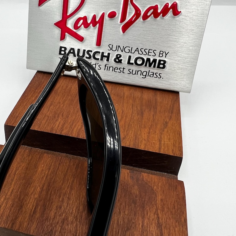 May include: Black Ray-Ban sunglasses with a brown lens, resting on a wooden display stand. The Ray-Ban logo is visible in red on a silver sign, with the text "SUNGLASSES BY BAUSCH & LOMB".