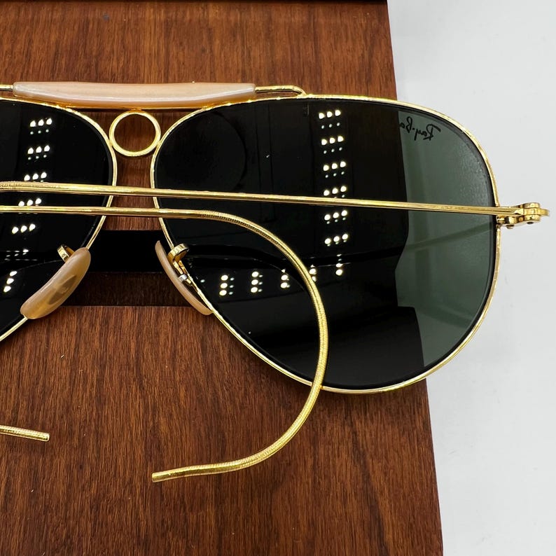 Pode incluir: &Oacute;culos de sol com arma&ccedil;&atilde;o dourada e lentes escuras. Os &oacute;culos t&ecirc;m uma ponte dupla e uma barra superior clara. As lentes s&atilde;o de cor verde escuro. Os &oacute;culos est&atilde;o sobre uma superf&iacute;cie de madeira.