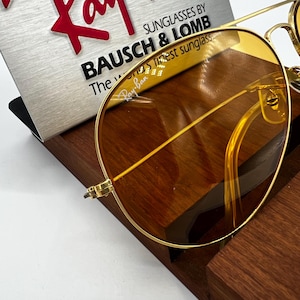 Puede incluir: Gafas de sol con montura de metal dorado y lentes tintadas en color &aacute;mbar. Las lentes presentan el logotipo de Ray-Ban en la esquina superior. Una tarjeta de presentaci&oacute;n detr&aacute;s de las gafas dice: Ray-Ban SUNGLASSES BY BAUSCH & LOMB The world's finest sunglasses.
