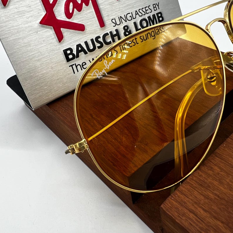 Puede incluir: Gafas de sol con montura de metal dorado y lentes tintadas en color &aacute;mbar. Las lentes presentan el logotipo de Ray-Ban en la esquina superior. Una tarjeta de presentaci&oacute;n detr&aacute;s de las gafas dice: Ray-Ban SUNGLASSES BY BAUSCH & LOMB The world's finest sunglasses.
