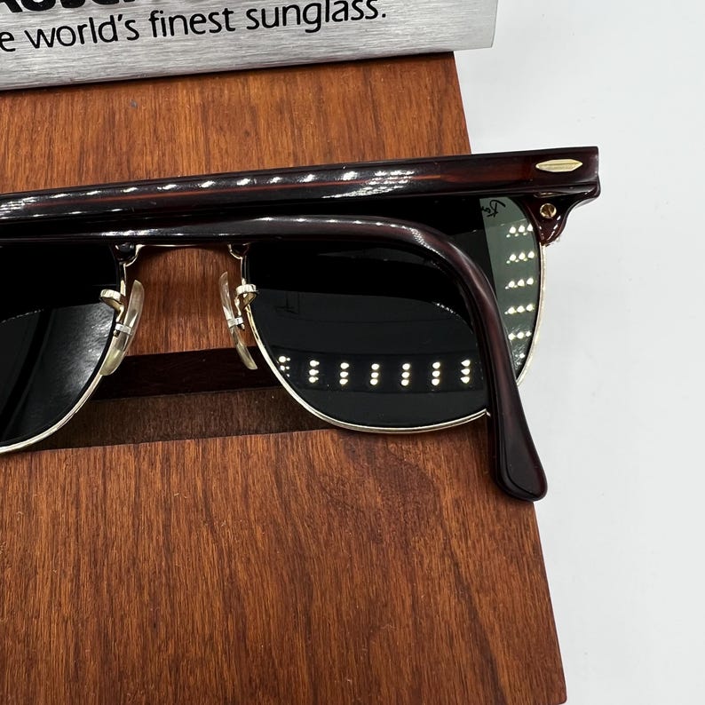May include: Close-up of sunglasses with dark lenses and a brown tortoiseshell frame. The sunglasses have gold-colored accents and are resting on a wooden surface. The text "world's finest sunglass" is visible.